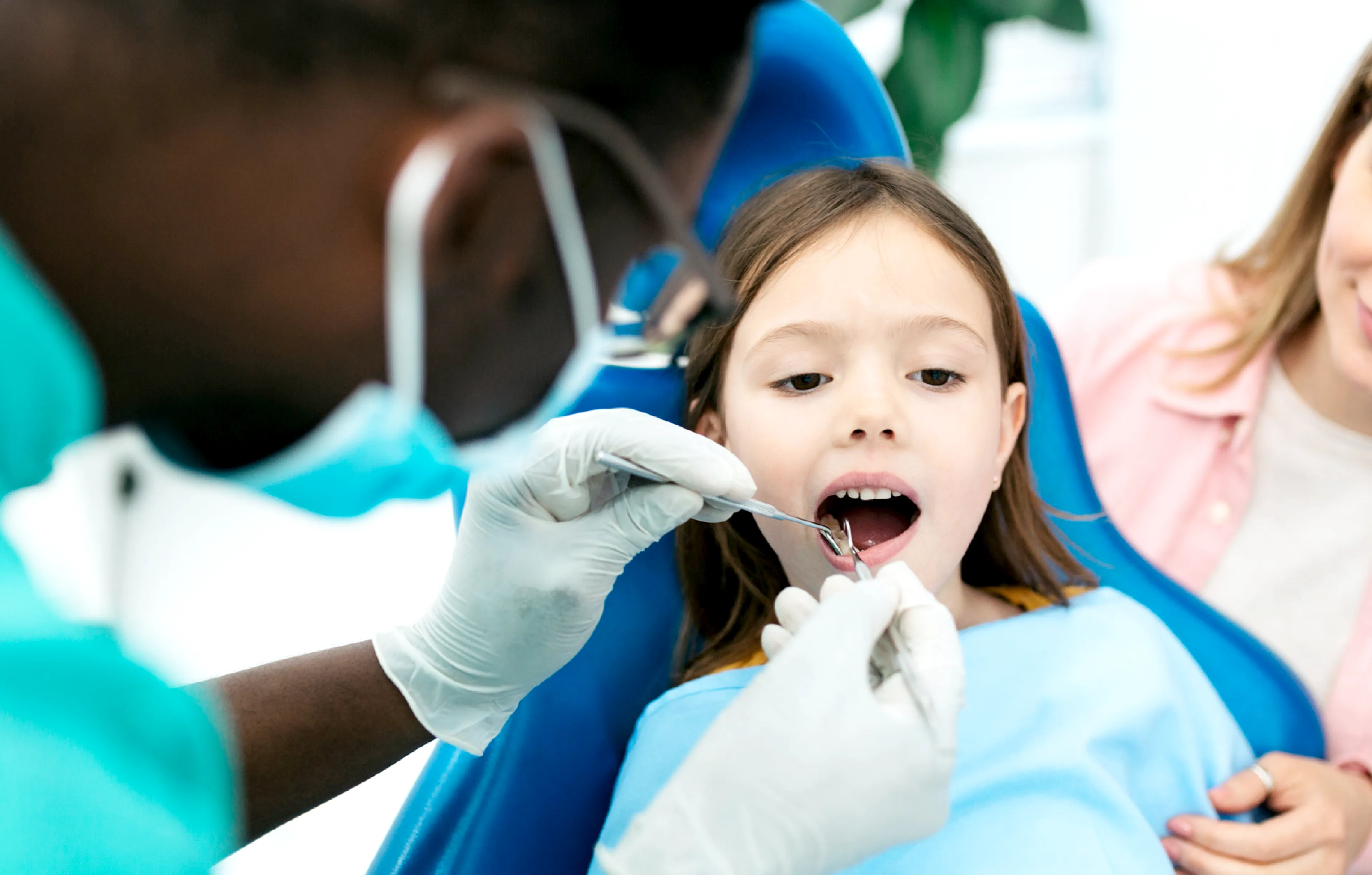 A young girl sits in a dental chair with her mouth open as a dentist examines her teeth using a dental mirror. A parent watches nearby.